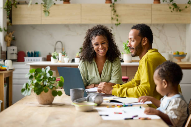 Freelancer discussing with smiling woman over laptop. Parents are sitting by toddler boy drawing at table. They are at home.